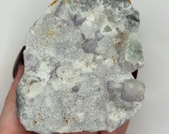 May include: A raw, natural fluorite specimen. The stone features a mix of colors, including pale purple, clear, and green crystals, set against a textured, gray matrix. The rough edges and varied surface create a unique, organic appearance.
