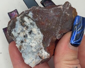 May include: A raw, irregular mineral specimen featuring a deep reddish-brown base with a prominent, textured patch of white, crystalline mineral deposits. The surface displays varied earthy tones and small, sparkling inclusions across its rough, jagged edges.