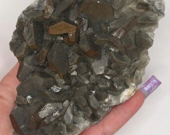 May include: A natural mineral specimen featuring a cluster of dark brown and gray crystals. The crystals have a geometric shape and are clustered together on a lighter gray base. The overall appearance is rough and textured.