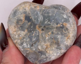 May include: A heart-shaped celestite crystal featuring a cluster of pale blue, translucent, and crystalline structures with visible tan and grey mineral inclusions throughout the textured, jagged surface of the stone.