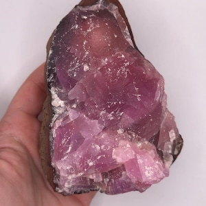 May include: A raw, uncut gemstone with a deep purple hue, transitioning to lighter pink shades. The stone has a rough, textured surface and is held in a hand against a white background. The edges are a dark brown color.