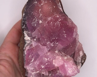 May include: A raw, uncut gemstone with a deep purple hue, transitioning to lighter pink shades. The stone has a rough, textured surface and is held in a hand against a white background. The edges are a dark brown color.