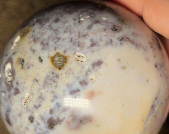 May include: A polished, spherical stone with a mix of colors. The surface displays a blend of creamy white, lavender, and gray hues, speckled with darker spots. The stone is held in a hand, showcasing its smooth texture and natural patterns.