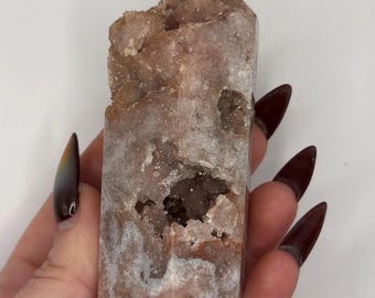 May include: A raw, tower-shaped crystal point with a dusty rose and brownish-tan coloration. The stone features a rough, textured surface with a prominent central vug or cavity filled with small, sparkling crystalline formations throughout.