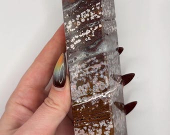 May include: A polished, tower-shaped crystal point featuring a deep brown and reddish-brown base color with distinct, scattered white floral-like inclusions throughout the stone. The surface is smooth and reflective with a pointed top.