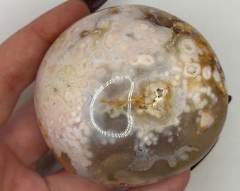 May include: A polished, rounded ocean jasper stone featuring a translucent, milky base with intricate, circular orbicular patterns in shades of cream, beige, soft pink, and light brown, with a small, distinct golden-brown mineral inclusion.