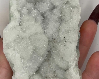May include: A light green and white crystal cluster with a sparkling, textured surface. The natural mineral formation has a rough, irregular shape and is held in a hand. The crystal appears to be a geode or druzy.