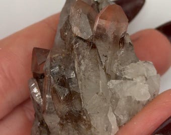 May include: A cluster of raw quartz crystals featuring translucent points with reddish-brown inclusions and dark smoky coloration at the base. The mineral specimen displays sharp, geometric terminations and a jagged, crystalline structure throughout.