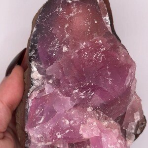 May include: A raw, uncut crystal specimen with a striking pink and purple hue. The mineral has a rough, textured surface with areas of crystalline structure. The edges are a darker brown color.