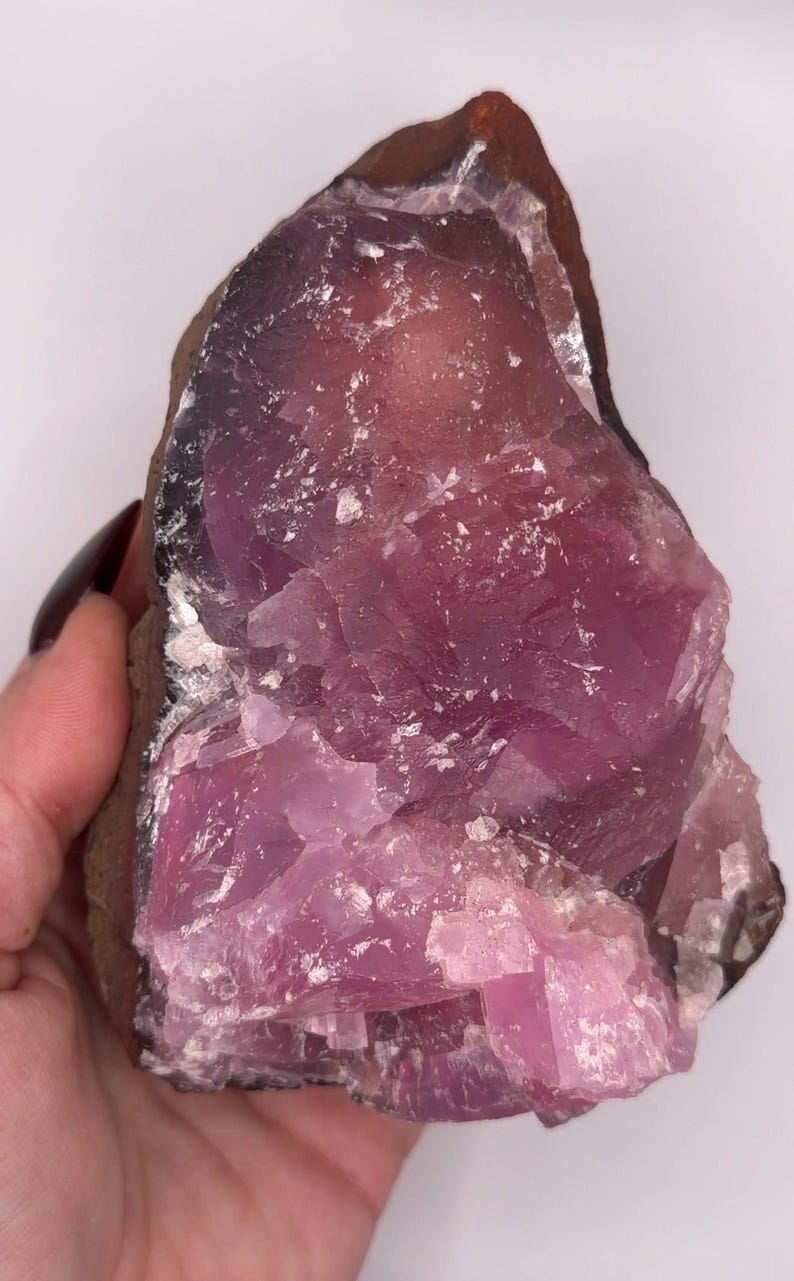May include: A raw, uncut crystal specimen with a striking pink and purple hue. The mineral has a rough, textured surface with areas of crystalline structure. The edges are a darker brown color.