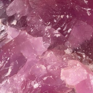 May include: Close-up of a raw, crystalline mineral with varying shades of pink and magenta. The rough, textured surface displays a mix of opaque and translucent areas, with small white inclusions.
