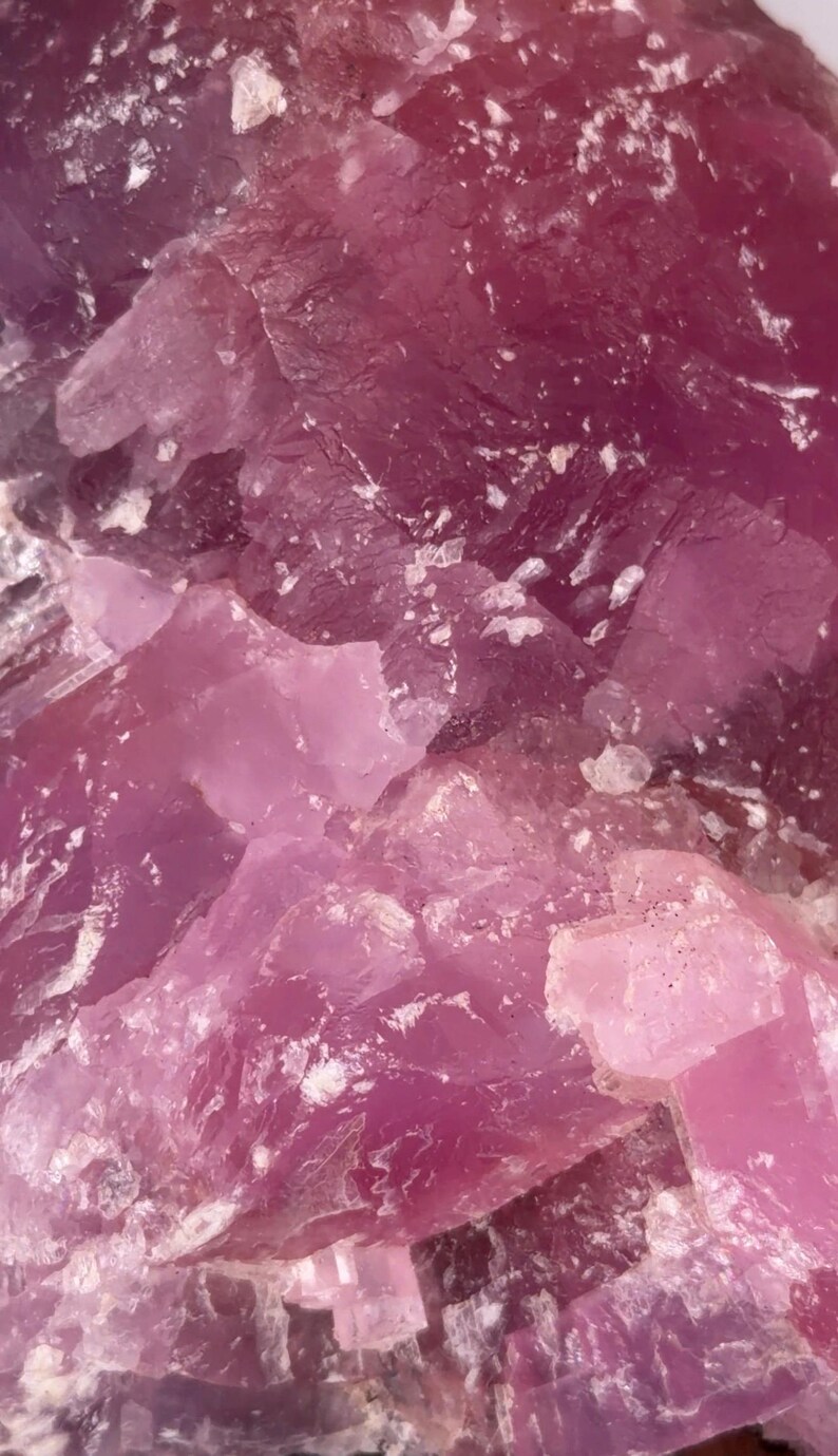 May include: Close-up of a raw, crystalline mineral with varying shades of pink and magenta. The rough, textured surface displays a mix of opaque and translucent areas, with small white inclusions.