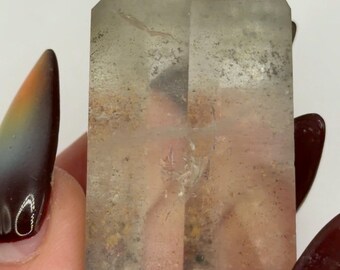 May include: A clear, six-sided quartz crystal point with a pointed top. The interior of the stone features visible inclusions of earthy brown and grey mineral deposits scattered throughout the translucent structure, creating a natural, textured appearance.