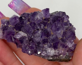 May include: A cluster of amethyst crystals, showcasing a deep purple hue with crystalline structures. The natural formation has a rough, textured surface and varying shades of violet. The crystals are held in a hand.