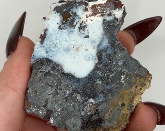 May include: A raw mineral specimen featuring a dark gray, metallic base with a prominent patch of opaque, milky white botryoidal mineral growth on the upper surface. The rock has an irregular, jagged shape with hints of brown and rust-colored oxidation.