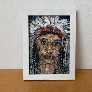 Puede incluir: Retrato de pintura al óleo de una persona nativa americana en un lienzo rectangular enmarcado en blanco. El retrato presenta a una persona que lleva un tocado con plumas blancas y una banda roja. La cara está pintada con varios colores.