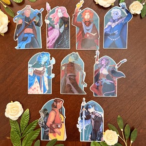 May include: A set of character stickers with fantasy art. The stickers show different figures in dynamic poses, with detailed clothing and accessories. The colour palette includes blues, reds, purples, and greens. Decorative floral elements surround the stickers.