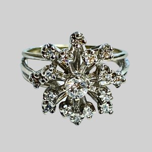Może przedstawiać: Srebrny pierścionek z dużym, okrągłym, czystym diamentem w środku. Diament jest otoczony mniejszymi diamentami, kt&oacute;re są osadzone w kwiatowym wzorze.