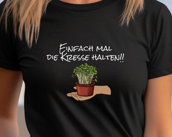 May include: Black t-shirt with a white graphic of a hand holding a small potted plant. The text on the shirt reads "Einfach mal die Kresse halten!"