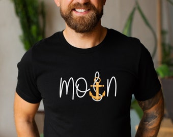Pode incluir: Camiseta preta com a palavra "mo&n" em escrita branca e um desenho de âncora dourada. A camiseta é de gola redonda e parece ser feita de um material macio. O desenho está centralizado no peito.