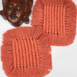May include: Two square, terracotta-colored macrame coasters with fringed edges. A carved wooden sculpture of a serene face is in the background. The coasters are made of woven cotton and are designed to protect surfaces from heat and moisture.