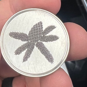 Puede incluir: Una moneda redonda de color plateado con un grabado detallado de una pi&ntilde;a rodeada de agujas de pino estilizadas. La moneda tiene un acabado de metal cepillado y un borde en relieve.