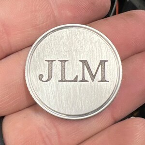 Puede incluir: Una moneda redonda de metal cepillado con las iniciales "JLM" grabadas en una fuente serif. La moneda tiene un borde elevado y una superficie texturizada, sostenida en una mano.