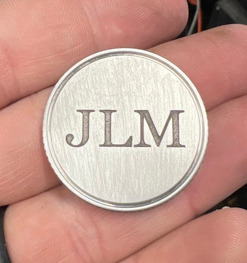 Puede incluir: Una moneda redonda de metal cepillado con las iniciales "JLM" grabadas en una fuente serif. La moneda tiene un borde elevado y una superficie texturizada, sostenida en una mano.