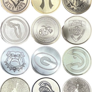 Puede incluir: Una colecci&oacute;n de monedas de metal plateado. Cada moneda presenta un dise&ntilde;o diferente, incluyendo logotipos de equipos deportivos, una puerta Torii japonesa, el emblema del Cuerpo de Marines de EE. UU. y el sello de la Universidad del Sur de California. Algunas monedas tienen texto como "TORONTO BLUE JAYS".