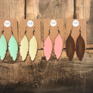 May include: Four pairs of leather feather earrings in different colors: mint green, yellow, pink, and brown. The earrings are displayed on a wooden background.