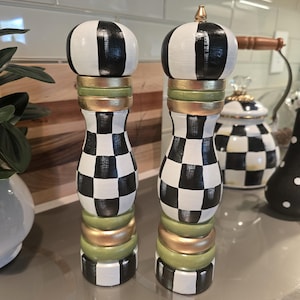 May include: Two tall salt and pepper grinders featuring a black and white checkered pattern on the main body and top. The cylindrical bodies are accented with horizontal bands of metallic gold and muted sage green paint, with a gold finial on top.