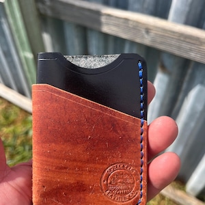 May include: A handcrafted leather card wallet with a brown exterior and black interior. The wallet features blue stitching along the edges and a stamped logo. The wallet is designed to hold cards and cash.