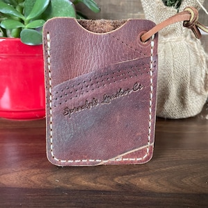 May include: A brown leather card holder with white stitching and a perforated design. The card holder has a small loop with a leather string attached. The text "Spreckels Leather Co." is embossed on the front.