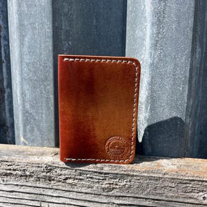 May include: A brown leather wallet with white stitching. The wallet has a rounded corner and a circular embossed logo on the front. The leather has a gradient color from light brown to dark brown.