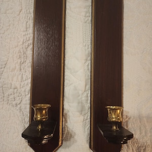 May include: Pair of vintage wall sconces. Each sconce features a dark brown wooden backplate with gold trim. A brass candle holder sits on a black base. The sconces are approximately 18 inches tall.