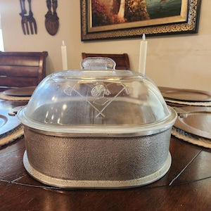 May include: An oval-shaped casserole dish with a textured, metallic gray base and a clear glass lid. The lid features an etched design of a knight's helmet centered between two crossed halberds, with a clear glass handle on top.