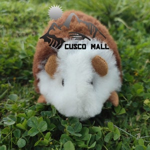 May include: A brown and white plush toy guinea pig with a brown nose and ears. The toy is sitting on a bed of green grass. The toy has the words "CUSCO MALL" printed on its back.