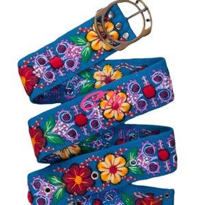 May include: A blue canvas belt with colorful embroidered flowers and a gold metal buckle. The belt has multiple eyelets for adjusting the fit.