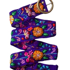 May include: A purple fabric belt featuring vibrant, multi-colored floral embroidery in shades of orange, pink, blue, and green. The belt includes a round, antique-style brass buckle and metal eyelets along the length of the strap for adjustment.