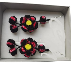 May include: A pair of black and red flower-shaped earrings with yellow centers, displayed in a white box.