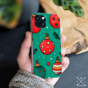 May include: A clear phone case featuring a festive Christmas ornament design. The case has a green background with red and green ornaments, white polka dots, and small sprigs of greenery. The ornaments have gold-colored tops and are in various styles.
