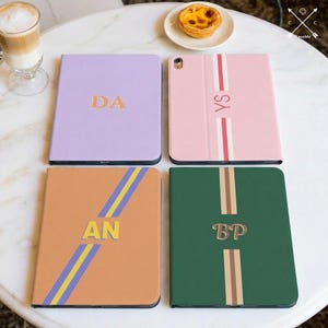 May include: Four tablet cases in various colors: lavender, pink, orange, and green. Each case features a unique design with initials and stripes. The lavender case has "DA", the pink case has "YS", the orange case has "AN", and the green case has "BP".