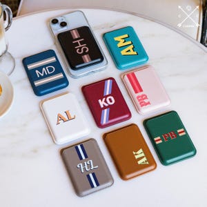 Könnte beinhalten: Eine Sammlung personalisierter Handy-Geldbörsen in verschiedenen Farben, darunter Blau, Türkis, Rot und Braun. Jede Geldbörse hat Initialen und Streifen in kontrastierenden Farben. Eine Geldbörse ist an einem Telefon befestigt.