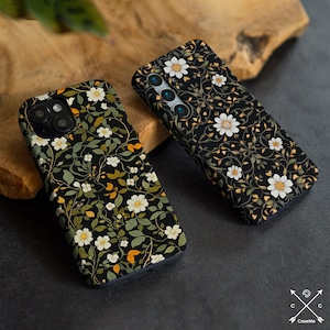 Op de afbeelding: Twee telefoonhoesjes met een bloemenpatroon. De hoesjes zijn zwart met witte bloemen en groene bladeren. De hoesjes liggen op een houten oppervlak.