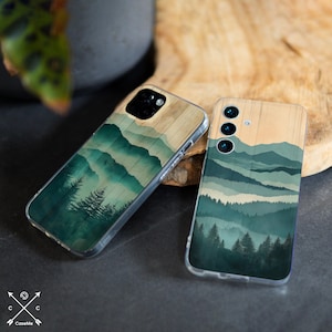 May include: Two phone cases with a nature-inspired design. The cases feature a wooden texture at the top and a landscape of green mountains and trees. The cases are clear and protect the phone.