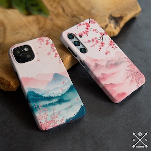 May include: Two phone cases with a watercolor design featuring pink and blue mountains, white clouds, and pink cherry blossoms. The cases are made of a hard plastic material and have a matte finish.