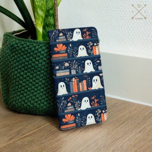 Pode incluir: Uma capa de telefone azul com um padrão de fantasmas brancos e livros laranja em uma estante. A capa tem um fundo azul escuro e os fantasmas são de diferentes tamanhos e formas.
