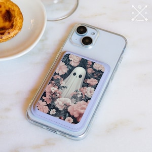 Pode incluir: Capa de telefone transparente com borda lavanda e design floral de fantasma. O design apresenta um fantasma branco rodeado por flores rosa e brancas sobre um fundo escuro. A capa está sobre uma superfície branca.