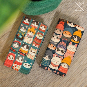 May include: Two teal blue wallet-sized phone cases with a repeating pattern of cats wearing winter hats and scarves. The cats are in various colors and patterns.