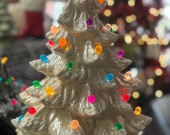 May include: Cream-colored ceramic Christmas tree with colorful lights and a red star topper. The tree has a textured surface and a gold leaf design at the base. A festive holiday decoration.